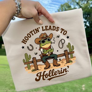 May include: Beige t-shirt featuring a cartoon frog dressed as a cowboy, wearing a hat and sunglasses, smoking a cigarette, and sitting on a wooden fence. The text "HOOTIN' LEADS TO HOLLERIN'" is printed above and below the frog.