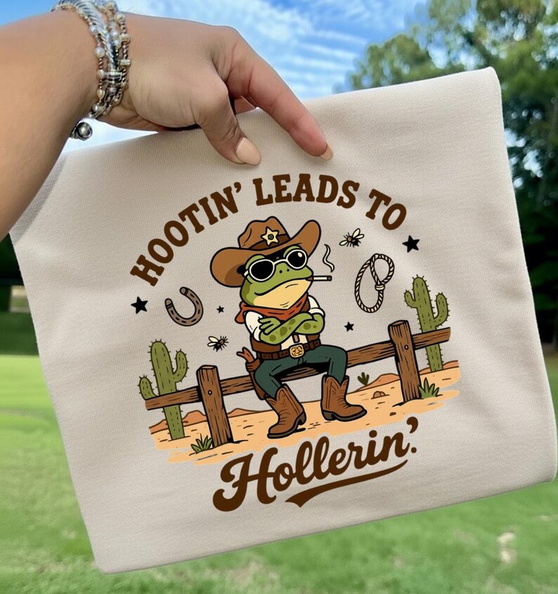 May include: Beige t-shirt featuring a cartoon frog dressed as a cowboy, wearing a hat and sunglasses, smoking a cigarette, and sitting on a wooden fence. The text "HOOTIN' LEADS TO HOLLERIN'" is printed above and below the frog.