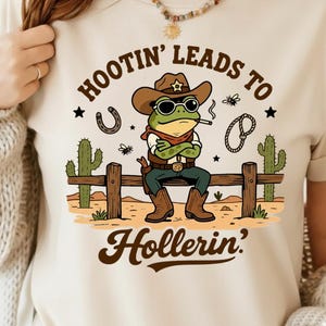 May include: Beige t-shirt with a cartoon frog cowboy graphic. The frog wears a brown hat, sunglasses, and a red bandana, smoking a cigarette. Text reads "Hootin' Leads to Hollerin'" with a horseshoe, rope, and cacti.
