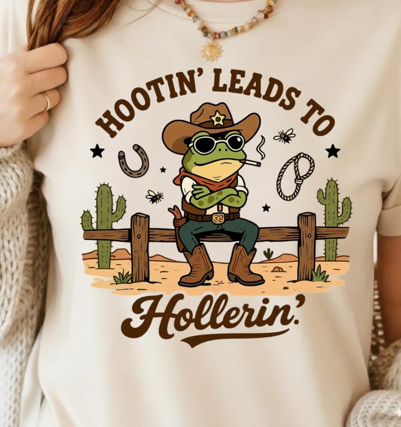 May include: Beige t-shirt with a cartoon frog cowboy graphic. The frog wears a brown hat, sunglasses, and a red bandana, smoking a cigarette. Text reads "Hootin' Leads to Hollerin'" with a horseshoe, rope, and cacti.