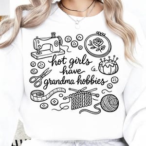 May include: White sweatshirt with a black graphic featuring sewing, embroidery, knitting, and other crafting elements. The text "hot girls have grandma hobbies" is written in a cursive font.