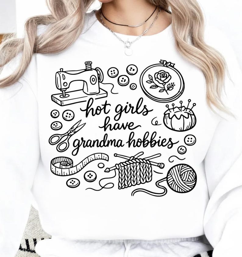 May include: White sweatshirt with a black graphic featuring sewing, embroidery, knitting, and other crafting elements. The text "hot girls have grandma hobbies" is written in a cursive font.