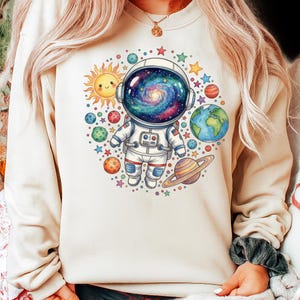 Peut inclure: Un sweat-shirt beige orn&eacute; d'une illustration color&eacute;e d'un astronaute avec un casque rempli de galaxies, entour&eacute; de plan&egrave;tes, d'&eacute;toiles et d'un soleil souriant. Le design a une esth&eacute;tique fantaisiste sur le th&egrave;me de l'espace.