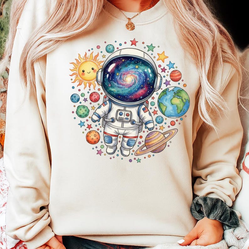 Peut inclure: Un sweat-shirt beige orn&eacute; d'une illustration color&eacute;e d'un astronaute avec un casque rempli de galaxies, entour&eacute; de plan&egrave;tes, d'&eacute;toiles et d'un soleil souriant. Le design a une esth&eacute;tique fantaisiste sur le th&egrave;me de l'espace.