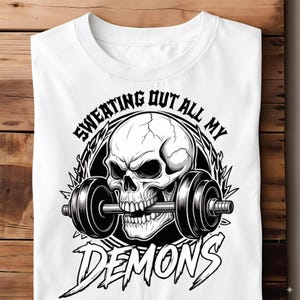 Może przedstawiać: Biała koszulka z czarno-białą grafiką czaszki trzymającej sztangę. Tekst "SWEATING OUT ALL MY DEMONS" jest wygięty nad i pod czaszką. Koszulka ma okrągły dekolt. Idealna na siłownię.