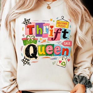 May include: Cream-colored sweatshirt with a colorful graphic that reads "Thrift Queen." The design includes a shopping basket, stars, and a "Second Hand" sign. The letters are in various colors.
