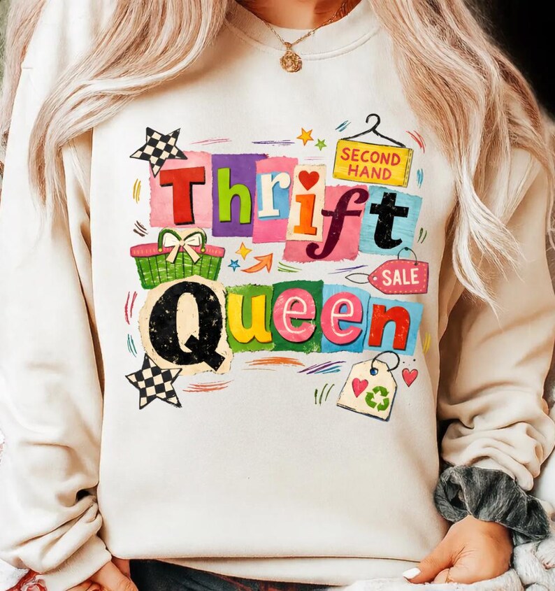 May include: Cream-colored sweatshirt with a colorful graphic that reads "Thrift Queen." The design includes a shopping basket, stars, and a "Second Hand" sign. The letters are in various colors.