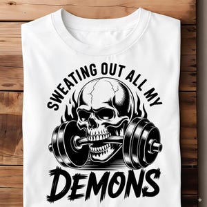 Może przedstawiać: Biała koszulka z czarno-białą grafiką czaszki podnoszącej sztangę. Tekst "SWEATING OUT ALL MY DEMONS" jest wydrukowany nad i pod wzorem czaszki i sztangi. Koszulka jest wykonana z miękkiego materiału.
