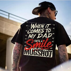 May include: Black t-shirt with the text "WHEN IT COMES TO MY DAD I WILL SMILE IN-MY MUGSHOT" in white and red. The shirt has a graphic design resembling a police lineup background. The person is wearing a black cap.