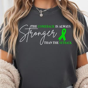 Könnte beinhalten: Dunkelgraues T-Shirt mit dem Text "The Comeback Is Always Stronger Than The Setback" in weißer Schrift. Eine grüne Schleife ist ebenfalls abgebildet. Das T-Shirt hat einen lässigen Stil.