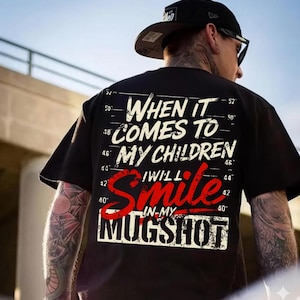 以下が含まれることがあります： 「WHEN IT COMES TO MY CHILDREN I WILL SMILE IN MY MUGSHOT」の文字が白と赤でプリントされた黒いTシャツ。かすれたフォントと測定スケールのグラフィックが特徴です。