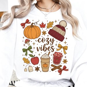 Könnte beinhalten: Weißes Sweatshirt mit einem herbstlichen Motiv. Das Design zeigt einen Kürbis, Blätter, eine Mütze und die Worte "cozy vibes". Weitere Elemente sind eine Kaffeetasse, ein Apfel und Zimtstangen.