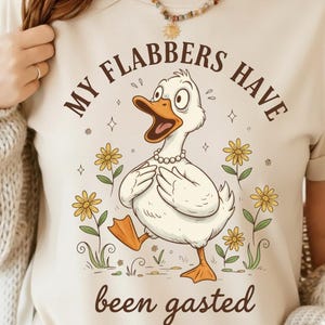 May include: Cream-colored t-shirt featuring a cartoon duck with an open beak, wearing a pearl necklace, and surrounded by yellow flowers. The text "MY FLABBERS HAVE been gasted" is arched above and below the duck.