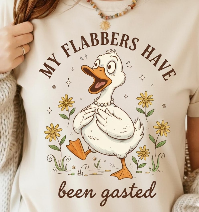 May include: Cream-colored t-shirt featuring a cartoon duck with an open beak, wearing a pearl necklace, and surrounded by yellow flowers. The text "MY FLABBERS HAVE been gasted" is arched above and below the duck.