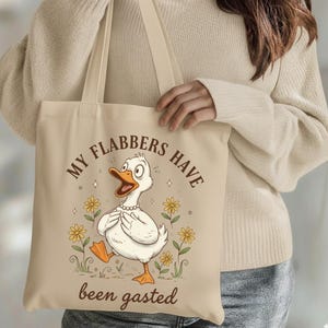 May include: Beige tote bag featuring a cartoon duck with the text "MY FLABBERS HAVE been gasted". The duck is white with an orange beak and feet, wearing a pearl necklace, and surrounded by yellow flowers. The bag has long beige straps.
