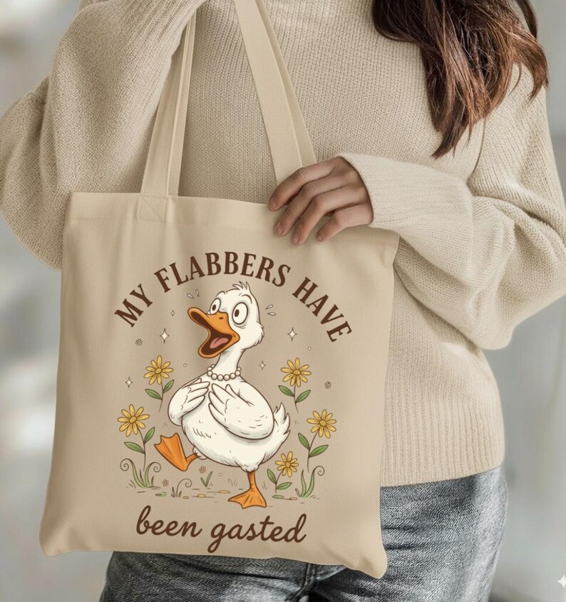 May include: Beige tote bag featuring a cartoon duck with the text "MY FLABBERS HAVE been gasted". The duck is white with an orange beak and feet, wearing a pearl necklace, and surrounded by yellow flowers. The bag has long beige straps.