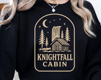 May include: Black sweatshirt with a beige graphic of a cabin, waterfall, trees, moon, and stars. The text "KNIGHTFALL CABIN" is below the cabin illustration. The sweatshirt is a crew neck style.