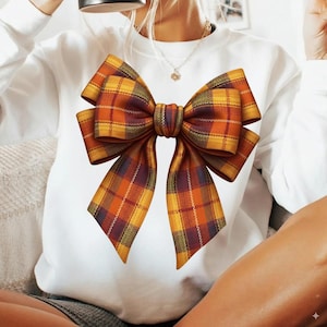 Könnte beinhalten: Weißes Sweatshirt mit einer großen Schleifen-Grafik. Die Schleife hat ein Karomuster in Orange-, Gelb- und Burgundertönen. Das Design ist mittig auf der Vorderseite des Kleidungsstücks platziert und erzeugt einen auffälligen visuellen Effekt. Das Muster der Schleife ist detailliert.