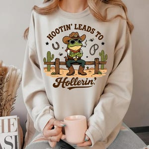 May include: Beige sweatshirt featuring a cartoon frog cowboy wearing sunglasses, a hat, and boots, smoking a cigarette. The text "Hootin' Leads to Hollerin'" is above and below the frog. A horseshoe, cactus, and other graphics are also included.