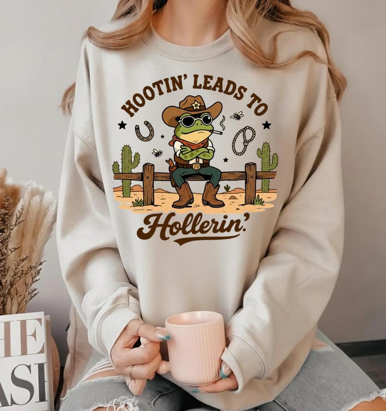 May include: Beige sweatshirt featuring a cartoon frog cowboy wearing sunglasses, a hat, and boots, smoking a cigarette. The text "Hootin' Leads to Hollerin'" is above and below the frog. A horseshoe, cactus, and other graphics are also included.