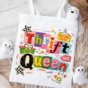 May include: White tote bag with the words "Thrift Queen" in colorful, block letters. The design includes a shopping basket, stars, hearts, and the words "Second Hand" and "Sale". Two small ghost-shaped plushies are visible.