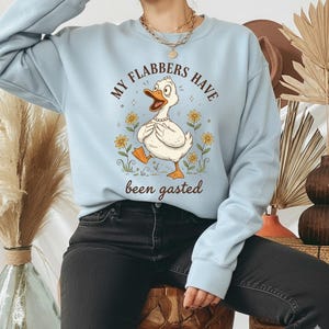 May include: Light blue sweatshirt featuring a cartoon duck with the text "MY FLABBERS HAVE been gasted." The duck is white with an orange beak and feet, wearing a pearl necklace, and surrounded by yellow flowers. The sweatshirt has long sleeves.