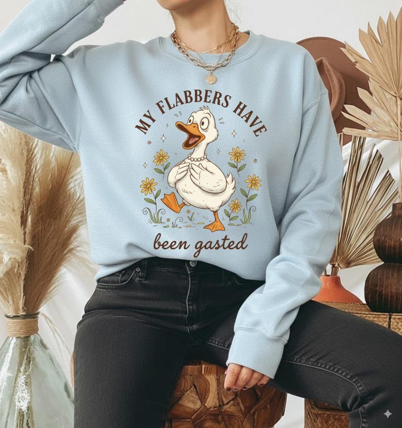 May include: Light blue sweatshirt featuring a cartoon duck with the text "MY FLABBERS HAVE been gasted." The duck is white with an orange beak and feet, wearing a pearl necklace, and surrounded by yellow flowers. The sweatshirt has long sleeves.