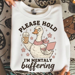 Op de afbeelding: Witte sweatshirt met een afbeelding van een cartoon eend met een ijskoffie en een roze laptop. De tekst luidt "PLEASE HOLD I'M MENTALLY buffering". De eend draagt een roze sjaal en is omringd door roze bloemen en sterren.