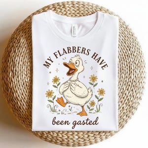 May include: White t-shirt with a cartoon duck graphic. The duck is wearing a pearl necklace and is surrounded by yellow flowers. Text on the shirt reads "MY FLABBERS HAVE been gasted."
