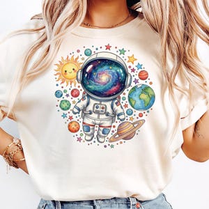 Peut inclure: T-shirt cr&egrave;me avec un astronaute de dessin anim&eacute; portant un casque rempli de galaxies, entour&eacute; de plan&egrave;tes, d'&eacute;toiles et d'un soleil souriant. L'astronaute porte une combinaison spatiale blanche avec des accents rouges et bleus. Design sur le th&egrave;me de l'espace.