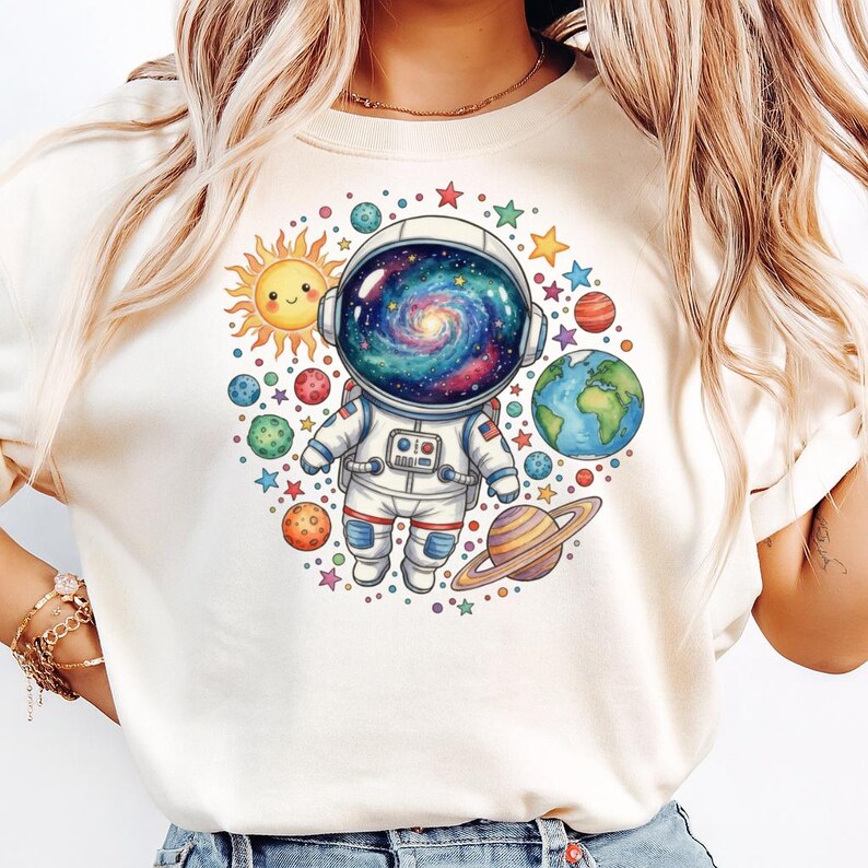 Peut inclure: T-shirt cr&egrave;me avec un astronaute de dessin anim&eacute; portant un casque rempli de galaxies, entour&eacute; de plan&egrave;tes, d'&eacute;toiles et d'un soleil souriant. L'astronaute porte une combinaison spatiale blanche avec des accents rouges et bleus. Design sur le th&egrave;me de l'espace.
