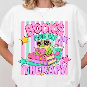 May include: White t-shirt featuring a colorful graphic. The design includes the text "BOOKS ARE MY THERAPY" in pink and green, a frog wearing heart-shaped sunglasses reading a book, and star and drink cup illustrations.