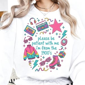 May include: White sweatshirt with a colorful 1900s-themed graphic. The design includes a boombox, cassette tape, sunglasses, a jacket, roller skate, and the text "please be patient with me I'm from the 1900's."