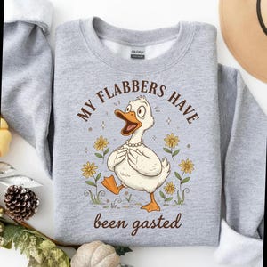 May include: A light gray sweatshirt featuring a cartoon duck with the text "MY FLABBERS HAVE been gasted". The duck is wearing a pearl necklace and surrounded by yellow flowers. The sweatshirt is a crew neck.