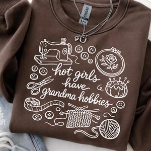 May include: A brown sweatshirt with white graphic illustrations of sewing, knitting, and embroidery tools. The text "hot girls have grandma hobbies" is also printed on the sweatshirt. A silver necklace is draped on the sweatshirt.