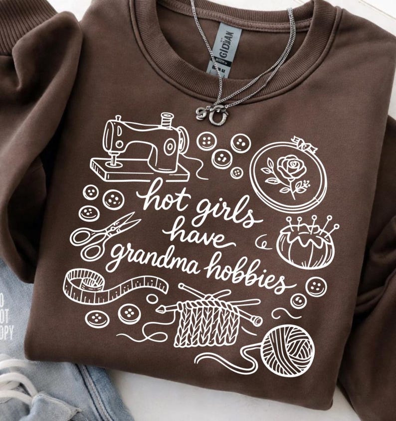 May include: A brown sweatshirt with white graphic illustrations of sewing, knitting, and embroidery tools. The text "hot girls have grandma hobbies" is also printed on the sweatshirt. A silver necklace is draped on the sweatshirt.