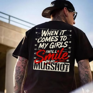 May include: Black t-shirt with the text "WHEN IT COMES TO MY GIRLS I WILL Smile IN-MY MUGSHOT" in white and red. The shirt has a graphic design resembling a police lineup background. The person in the image is wearing a black cap and sunglasses.