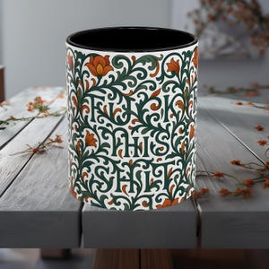 May include: A ceramic coffee mug with a black interior and a white exterior featuring a repeating pattern of green vines, orange flowers, and the words "F**K THIS SH*T" in green lettering.