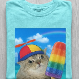 May include: A light blue t-shirt featuring a graphic of a cat wearing a multi-colored propeller hat, holding a rainbow-colored popsicle against a blue sky background with a rainbow. The inner neck label displays the word COMFORT in black text.