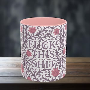 May include: A ceramic mug with a pink interior and a white exterior adorned with a repeating floral pattern in purple and pink. The words "FUCK THIS SHIT" are centered on the mug in a stylized font.