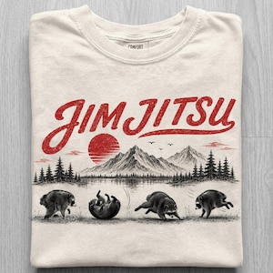 May include: A cream-colored t-shirt featuring a red graphic with the text "JIM JITSU" above a black and white illustration of mountains, a sun, and four raccoons in various martial arts poses on a grassy field with pine trees.