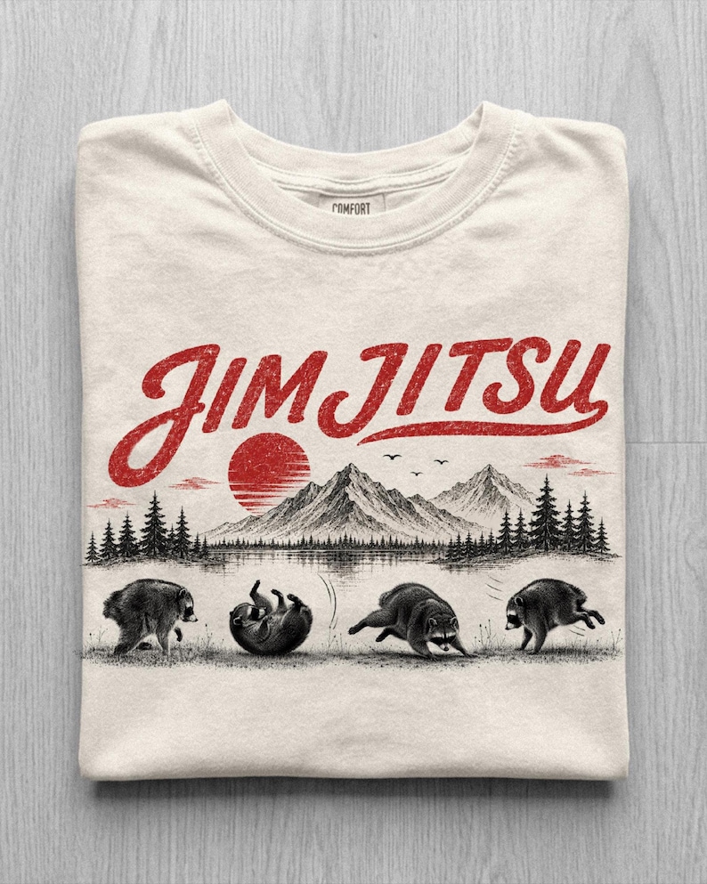 May include: A cream-colored t-shirt featuring a red graphic with the text "JIM JITSU" above a black and white illustration of mountains, a sun, and four raccoons in various martial arts poses on a grassy field with pine trees.