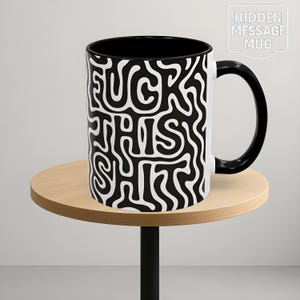May include: A white ceramic mug with a black interior and handle. The mug features a black and white abstract design with the words "FUCK THIS SHIT" in a bold, stylized font. The mug is on a small, round wooden table.