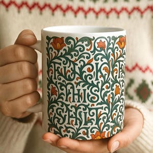 May include: White ceramic mug with a green, orange, and white floral design and the text "F**k This S**t". The mug is held by a person wearing a cream-colored sweater with a red and green pattern.