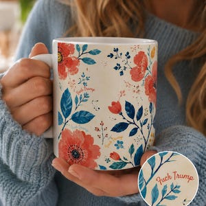 May include: White ceramic mug with a floral design featuring red and orange flowers, blue leaves, and the text "Fuck Trump". The mug has a white handle and is held by a person wearing a blue sweater.