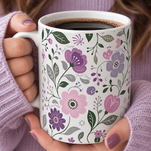 May include: White ceramic mug filled with dark coffee, featuring a floral pattern in shades of purple and pink, with green leaves and the text "Fuck Trump" repeated throughout the design.