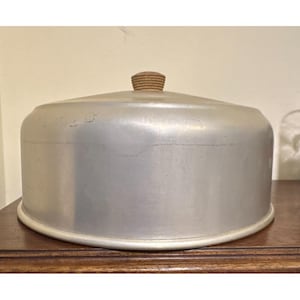 Puede incluir: Una cubierta para pasteles redonda de aluminio cepillado con una parte superior abovedada lisa y un borde de base ligeramente ensanchado. La cubierta cuenta con un pequeño pomo circular de madera clara centrado en la parte superior.