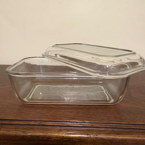 May include: A clear rectangular glass baking dish with a matching glass lid. The dish features a textured base with horizontal lines and a smooth rim. The lid is also rectangular with a raised center section and a smooth, clear glass finish.