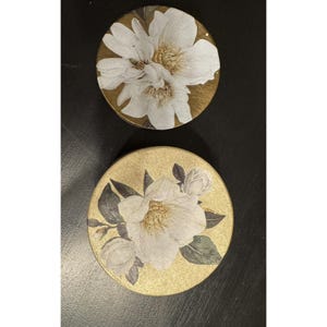 Op de afbeelding: Twee ronde onderzetters met een goudkleurige achtergrond en witte bloemillustraties. De bovenste onderzetter toont een grote witte bloem, terwijl de onderste onderzetter een witte bloem met groene bladeren en kleine knoppen in een gedetailleerd ontwerp laat zien.