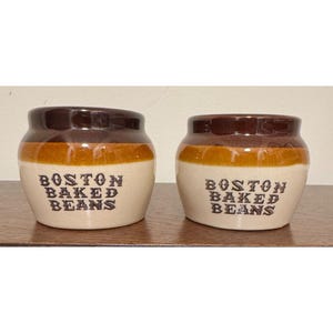 May include: Two ceramic bean pots featuring a two-tone glaze with a dark brown rim and a cream-coloured base. Each pot is printed with the black text BOSTON BAKED BEANS on the front. The pots have a rounded shape with a wide opening at the top.